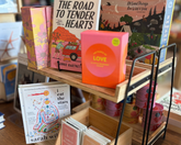 A wooden and black metal display stand holding assorted books, a bright pink box with a yellow 'LOVE' label, and small cards or booklets on a wooden table in a bookstore.