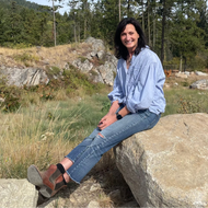 A woman sitting on a large rock in an outdoor mountain setting with pine trees and dry grass, wearing a light blue button-up shirt, blue jeans, and brown shoes.
