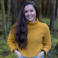 A woman with long dark hair smiling in a forest, wearing a mustard yellow ribbed turtleneck sweater and light blue jeans.