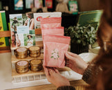 A person holds a stack of pink pouches with a white flower illustration in a cozy, book-filled shop setting.