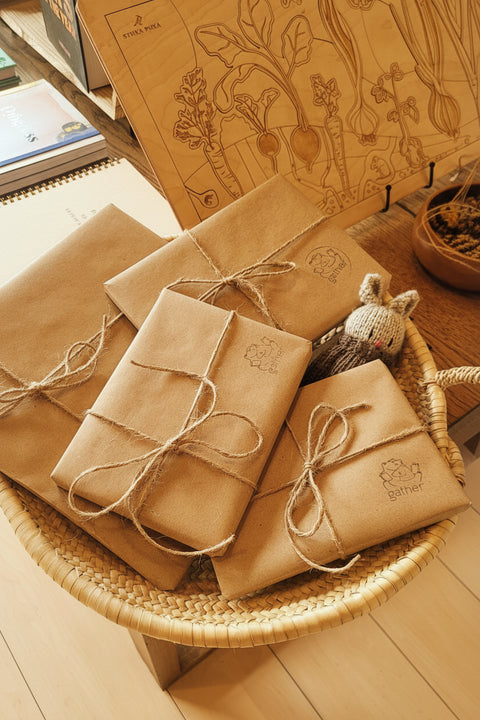 Three kraft paper gift boxes tied with twine bows and decorated with floral stamps, arranged in a woven basket on a wooden floor next to a wooden box with engraved botanical illustrations.