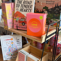 A wooden and black metal display stand holding assorted books, a bright pink box with a yellow 'LOVE' label, and small cards or booklets on a wooden table in a bookstore.
