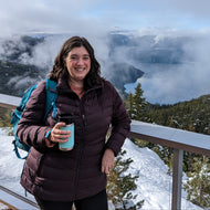 A woman wearing a dark plum puffer jacket and a teal backpack stands on a snowy mountain overlook holding a light blue tumbler, with a dramatic mountain lake and forest landscape in the background under a cloudy sky.