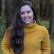 A smiling young woman with long dark brown hair wearing a mustard yellow ribbed turtleneck sweater, photographed outdoors against a blurred forest background.