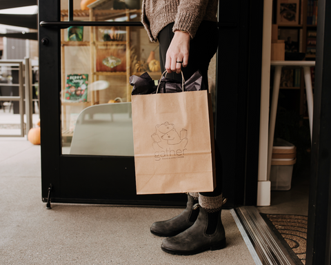 A person standing at a storefront doorway holding a brown paper shopping bag with a simple line drawing logo and the word 'gather' printed on it. The person is wearing a beige sweater, black pants, and dark gray ankle boots.