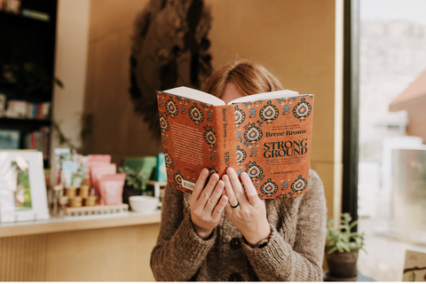 A person in a brown knit sweater holds a book titled 'Strong Ground' by Brené Brown in front of their face in a warmly lit indoor setting.